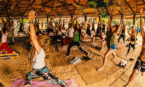 Grupo de pessoas praticando yoga em posições de guerreiro dentro de um domo de bambu com teto decorado, durante um festival na Aldeia Outro Mundo.
