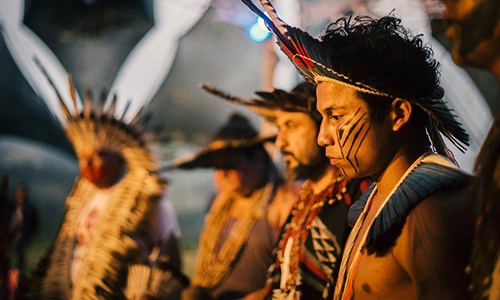 Participantes indígenas com pintura facial e cocares em uma cerimônia espiritual, representando a cultura de paz no festival Gaia Connection da Aldeia Outro Mundo.