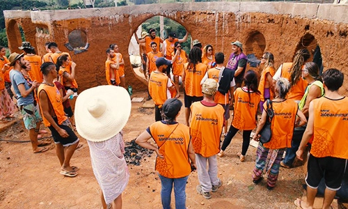Grupo de participantes em um workshop de bioconstrução, usando camisetas laranja com logo da Aldeia Outro Mundo, aprendendo técnicas de construção sustentável.