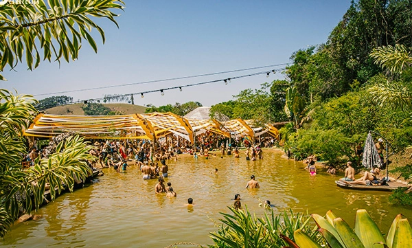 Pessoas se refrescando em uma piscina natural durante o festival Gaia Connection, com tendas e decoração orgânica na Aldeia Outro Mundo.