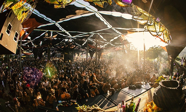 Multidão de pessoas dançando e celebrando no palco principal do festival Mundo de Oz, com luzes e estruturas decoradas na Aldeia Outro Mundo.